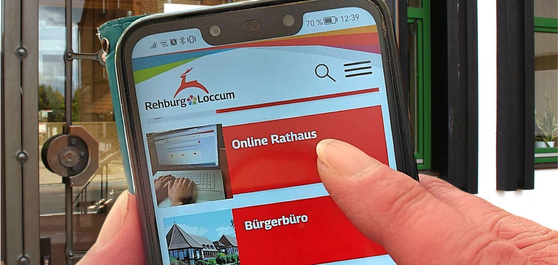 Auf einem Smartphone sieht man den Menüpunkt Online Rathaus und im Hintergrund den Eingang des Rathauses in Rehburg-Loccum