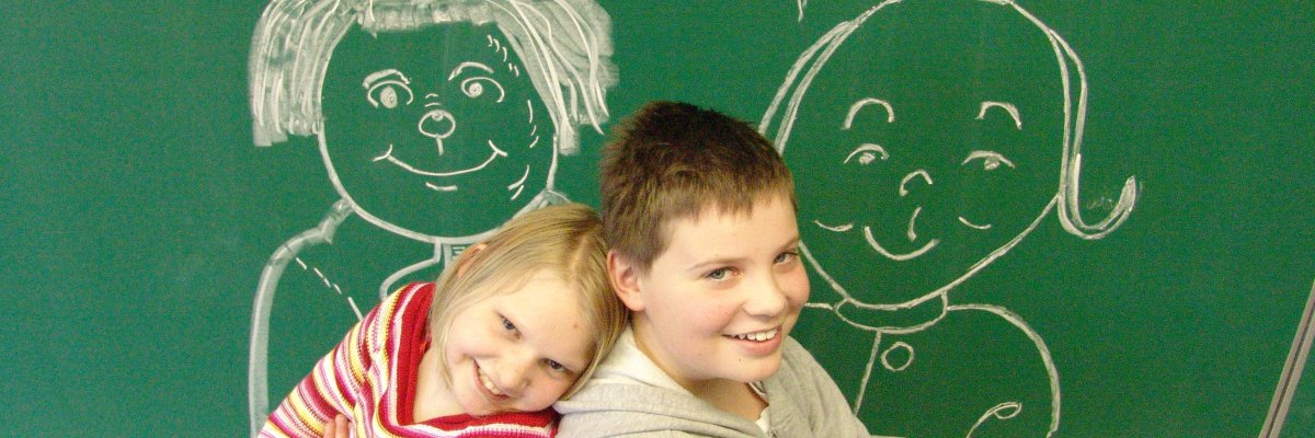 Ein Mädchen und ein Junge im Grundschulalter stehen vor einer grünen Schultafel, auf die die Max- und Moritz-Figuren gezeichnet sind, und lachen fröhlich.