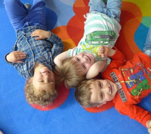 3 Kinder liegen auf dem Rücken auf einem bunten Teppich und lachen von unten in die Kamera