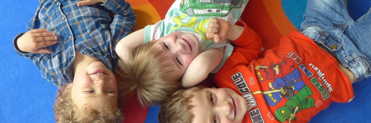 3 Kinder liegen auf dem Rücken auf einem bunten Teppich und lachen von unten in die Kamera
