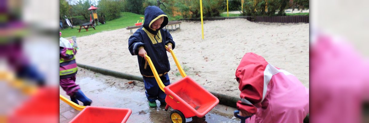 Wasserspiele 3 kleine Kinder in bunter Regenkleidung spielen im regen mit roten Schubkarren