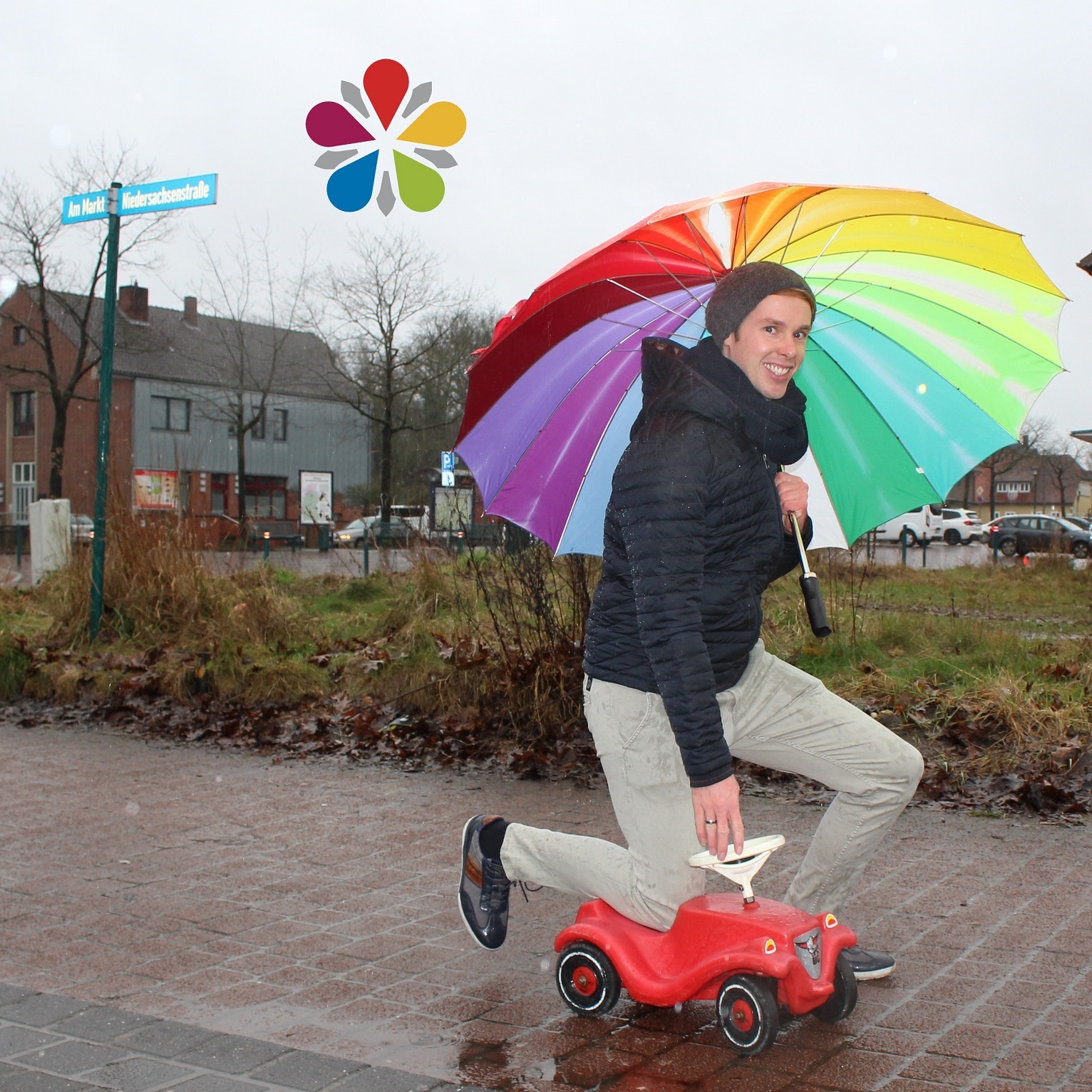 Sascha Rode kniend auf einem rotem Bobbycar mit einem buntem Regenschirm in  seiner Hand
