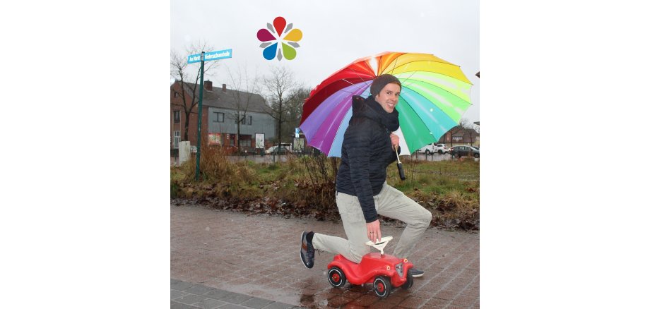 Sascha Rode kniend auf einem rotem Bobbycar mit einem buntem Regenschirm in  seiner Hand